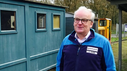 Hannsjörg Frank, lichtes weißes Haar, schwarze Brille, blaue Jacke, links dahinter sieht man die orangene Muttenthalbahn