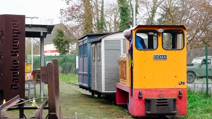 Blick von hinten auf die Muttenthalbahn, bestehend aus einer orangenen Lok, in der Hansjörg Frank steht und seitlich rausguckt, und einem grauen und einem blauen Waggon, die über die Gleise fährt