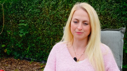 Julia Korbach auf einem Stuhl im Garten. Sie trägt einen rosa-farbenen Pullover.