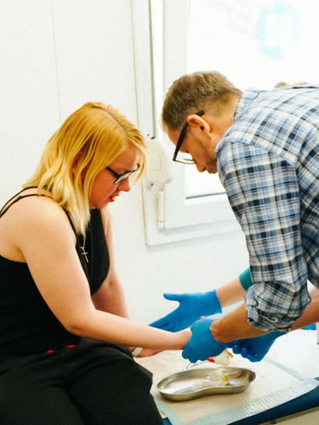 Carlo Vivalditrägt eine Brille, hat kurze braun-graue Haare, ein blau-weiß-kariertes Hemd, trägt blaue Handschuhe aus Gummi und hält die Hand einer Patientin mit Brille, blonden Haaren und schwarzem Tank-Top.