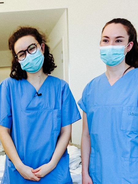 Lara Soyubey und Xenia Look stehen gemeinsam vor einer Patientin. Die beiden jungen Frauen tragen beide einen Mundschutz.
