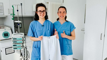 Lara Soyubey und Xenia Look lächeln in die Kamera, Lara Soyubey hält eine weiße Hose in die Luft.