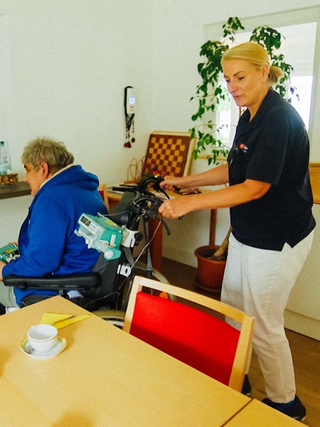 Krankenschwester Manuela Suler schiebt einen Hospiz-Patienten im Rollstuhl an einen Tisch.