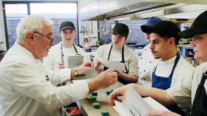 Ausbilder Manuel Klinger steht mit mehreren Azubis an einem Tisch in der Küche.
