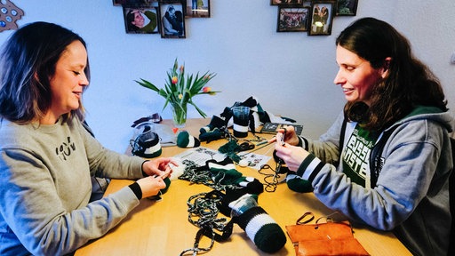 Zwei Frauen sitzen sich an einem Tisch gegenüber und häkeln. Zwischen ihnen liegt schwarz-weiß-grüne Häkelwolle.