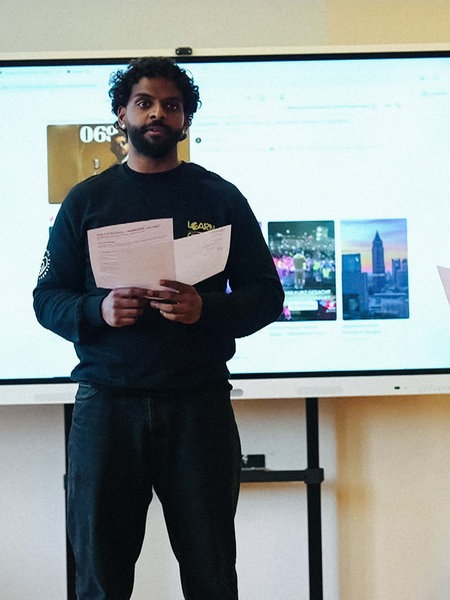 Zwei Männer, Kevin Grund und Nigash Sriranganathan, stehen mit Zetteln in der Hand in einer Schulklasse, hinter ihnen ist eine Präsentation zu sehen.