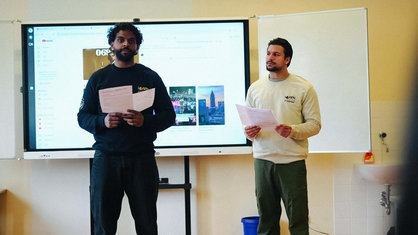 Zwei Männer, Kevin Grund und Nigash Sriranganathan, stehen mit Zetteln in der Hand in einer Schulklasse, hinter ihnen ist eine Präsentation zu sehen.