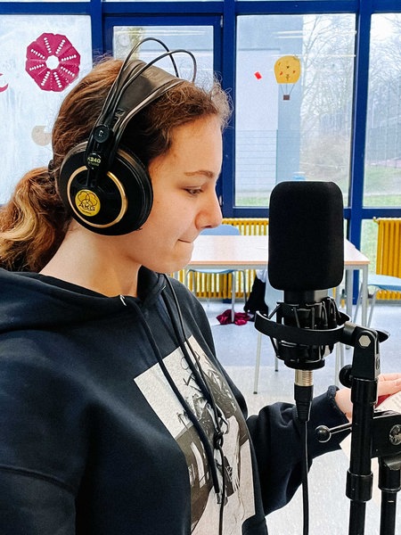 Cheyenne Erler steht beim Rap-Workshop im Klassenzimmer mit Kopfhörern vor einem Mikrofon. In der Hand hat sie einen Zettel mit ihrem Text.
