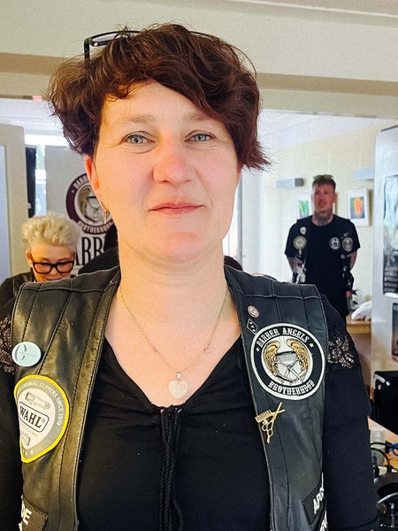 Eine Frau mit kurzen Haaren schaut in die Kamera. Sie trägt eine schwarze Weste mit bunten Stickern. Im Hintergrund werden Haare geschnitten.