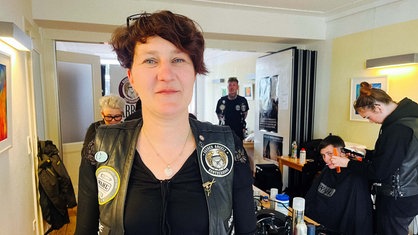 Eine Frau mit kurzen Haaren schaut in die Kamera. Sie trägt eine schwarze Weste mit bunten Stickern. Im Hintergrund werden Haare geschnitten.