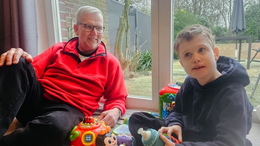 Ein Mann mit grauen Haaren und rotem Pullover auf einer Decke mit Spielzeug. Neben ihm ein kleinerer Junge mit dunkelblonden Haaren