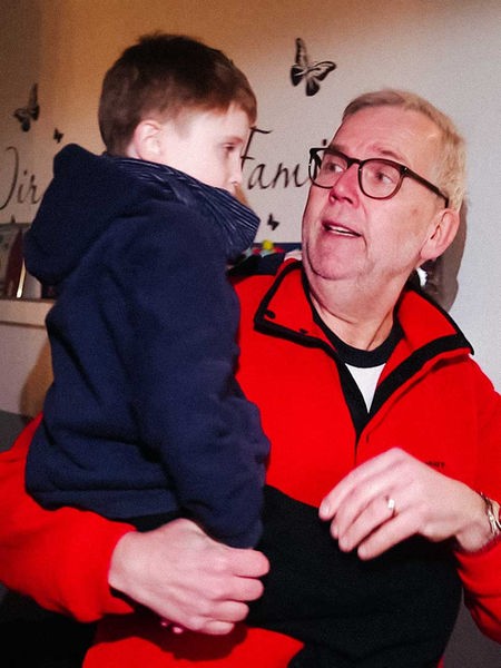 Ein Mann mit grauen Haaren und rotem Pullover auf einer Decke mit Spielzeug. Neben ihm ein kleinerer Junge mit dunkelblonden Haaren