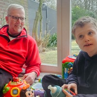 Ein Mann mit grauen Haaren und rotem Pullover auf einer Decke mit Spielzeug. Neben ihm ein kleinerer Junge mit dunkelblonden Haaren