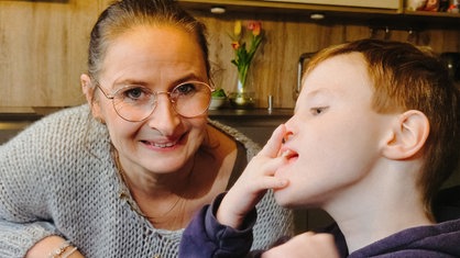 Eine Frau mit dunkelbraunem Zopf und goldener Brille. Neben ihr ein kleiner Junge mit blonden Haaren.
