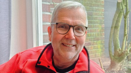 Ein Mann mit kurzen grauen Haaren und Brille. Er trägt einen roten Pullover.