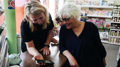 Vera Wipprecht (rechts) und eine Kollegin stehen an der Kasse im Dorfladen. Sie schauen auf ein Handy.