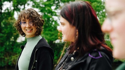 Drei Frauen gehen im Park spazieren. Die Frau ganz links ist im Fokus. Sie hat braune, lockige Haare und trägt eine Brille.