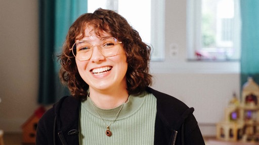 Eine junge Frau mit Brille, braunen, lockigen Haaren lächelt in die Kamera.