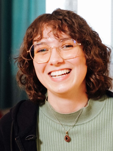 Eine junge Frau mit Brille, braunen, lockigen Haaren lächelt in die Kamera.