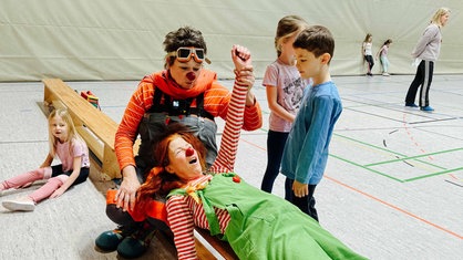 Die Clowns liegen auf einer Bank in der Turnhalle und albern mit den Schülern herum