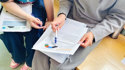 Zwei Schülerinnen sitzen auf einer Bank und haben jeweils einen Collegeblock und ein Papier auf ihrem Schoß. Sie halten einen Stift in ihrer Hand.