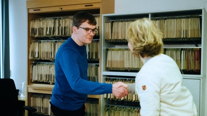 Andreas Kraschl schüttelt einer Frau die Hand. Sie stehen in einem Büro, wo in Regalen an der Wand unzählige Akten einsortiert sind.