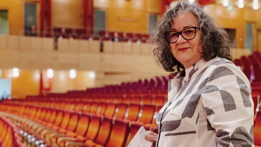 Silvia steht vor einer Sitzreihe der Essener Philarmonie. Das gesamte Gebäude ist menschenleer. In der rechten Hand hält sie ein ausgedrucktes Ticket. 