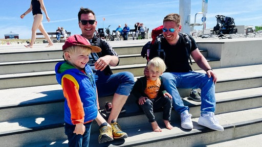 Die zwei Männer Matthias Apken und Matthias Vollmer-Lentmann und zwei kleine Jungen sitzen auf einer Treppe am Strand.