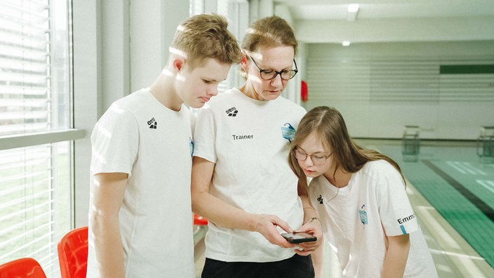 Sybille Vorwerk hält ein Handy in den Händen. Felix und Emma stehen neben ihr und schauen auf das Display.