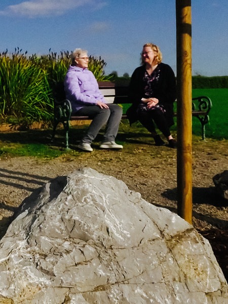 Zwei Frauen sitzen auf einem Friedhof auf einer Bank, im Vordergrund eine Engelsfigur