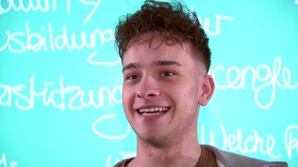 Janic Dalwigk, kurze braune Haare, die ihm über die Stirn fallen, vor einer hellblauen Wand, auf der weiße Schrift zu sehen ist