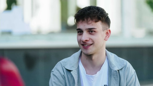 Janic Dalwigk, kurze braune Haare, die ihm über die Stirn fallen, grau-gestreiftes Hemd, darunter ein weißes T-Shirt.