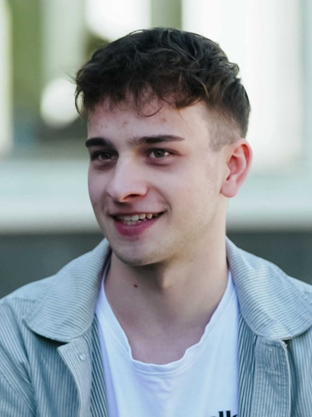 Janic Dalwigk, kurze braune Haare, die ihm über die Stirn fallen, grau-gestreiftes Hemd, darunter ein weißes T-Shirt.