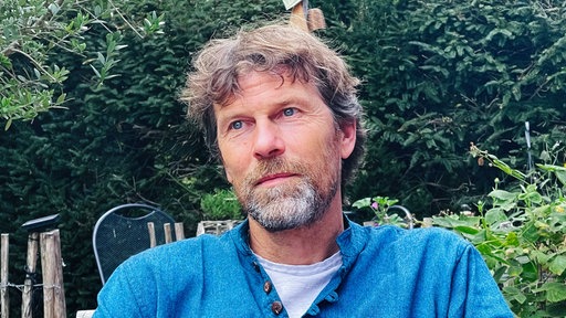 Ein Mann, Matthias Bäcker, in einem blauer Pullover sitzt in einem Garten.
