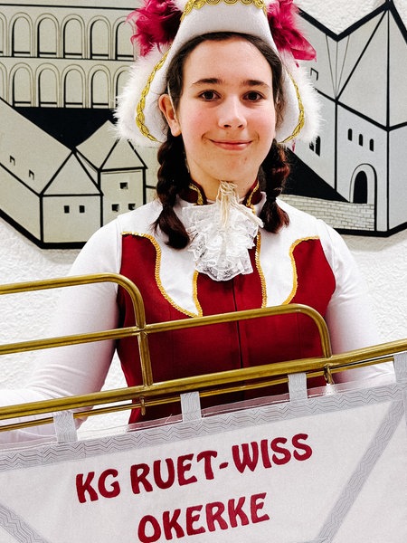 Vanessa Buch steht vor einer Wand an der eine Zeichnung einer Burg und das Wappen von Mönchengladbach hängt. In den Händen hält sie einen Banner mit der Aufschrift "KG Ruet-Wiss-Okerke". 