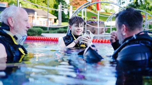 Jannick steht im Schwimmbecken. Er hält eine Taucherbrille in den Händen und lächelt. An den Rändern des Bildes schwimmen zwei Tauchassistenten.