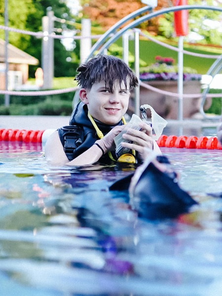 Jannick steht im Schwimmbecken. Er hält eine Taucherbrille in den Händen und lächelt. An den Rändern des Bildes schwimmen zwei Tauchassistenten.