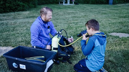 Luis (rechts) und sein Tauchbuddy (links) sitzen auf einer Wiese. Luis übt den Umgang mit der Sauerstoffflasche.