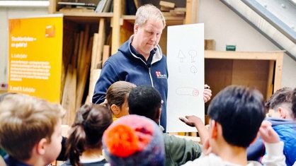 Tischlermeister Tobias Gregor hält ein Bild mit verschiedenen Zeichnungen in der Hand. Vor ihm steht eine Gruppe Schüler.