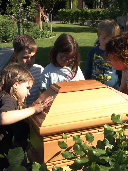 Pädagogin Karin Gerdemann und vier Kita-Kinder stehen auf der Trauerinsel auf dem Friedhof in Greven um das abgeschnittene Ende eines Sarges herum