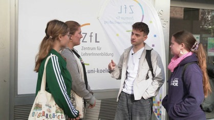 Janic Dalwigk steht vor einem weißen Banner, darauf steht "Zentrum für LerherInnenausbildung", rechts und links von ihm stehen drei junge Frauen, die ihn angucken, er gestikuliert mit einer Hand