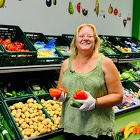 Eine Frau hält Gemüse in den Händen und posiert vor Kisten voller Gemüse und Kartoffeln.
