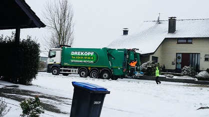 Ein Müllwagen mit einem Müllwerker hinten drauf und einem Müllwerker, der hinterher läuft, fährt eine verschneeite Straße entlang.