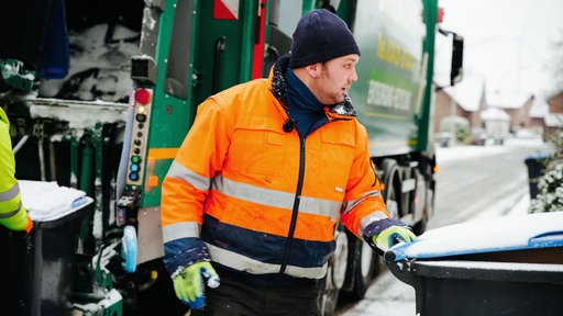 Martin Houtbeckers, ein Mann in orangener Jacke, steht neben einem Müllwagen, seine Hand liegt am Griff einer Papiertonne.