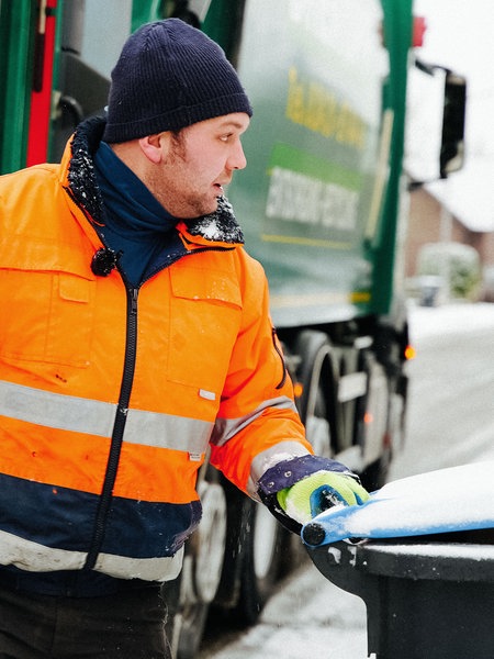Martin Houtbeckers, ein Mann in orangener Jacke, steht neben einem Müllwagen, seine Hand liegt am Griff einer Papiertonne.