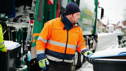 Martin Houtbeckers, ein Mann in orangener Jacke, steht neben einem Müllwagen, seine Hand liegt am Griff einer Papiertonne.