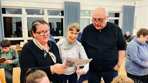 Eine Frau mit Behinderung steht lachend zwischen einem Mann und einer Frau mit dunklen Oberteilen, die ein Liedblatt in der Hand halten