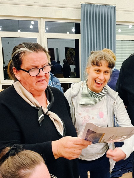 Eine Frau mit Behinderung steht lachend zwischen einem Mann und einer Frau mit dunklen Oberteilen, die ein Liedblatt in der Hand halten