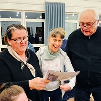 Eine Frau mit Behinderung steht lachend zwischen einem Mann und einer Frau mit dunklen Oberteilen, die ein Liedblatt in der Hand halten