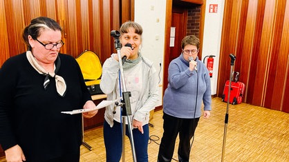 Eine Frau miit Behinderung steht lachend an einem Mikrofon. Neben ihr zwei Frauen, die ebenfalls singen
